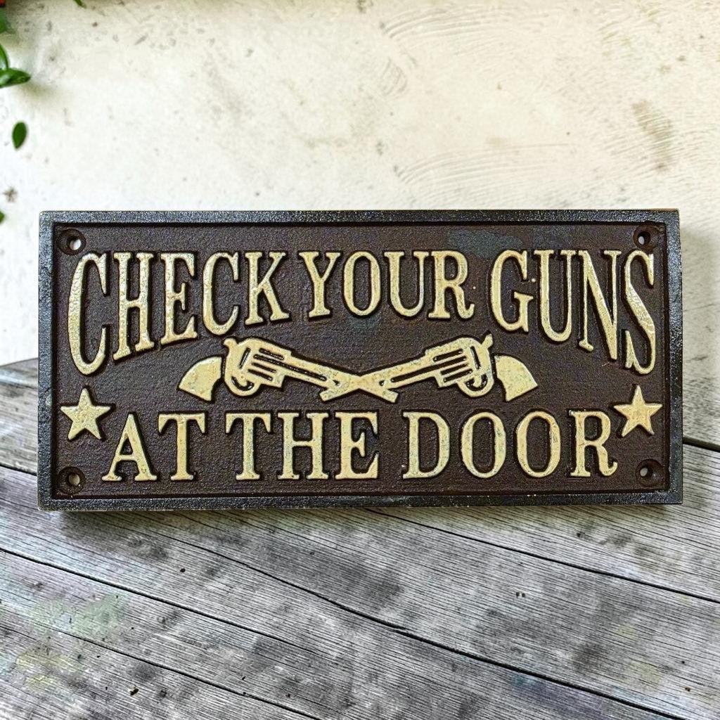 Jones Peak cast iron wall plaque with raised text "Check Your Guns At The Door" and crossed revolvers.